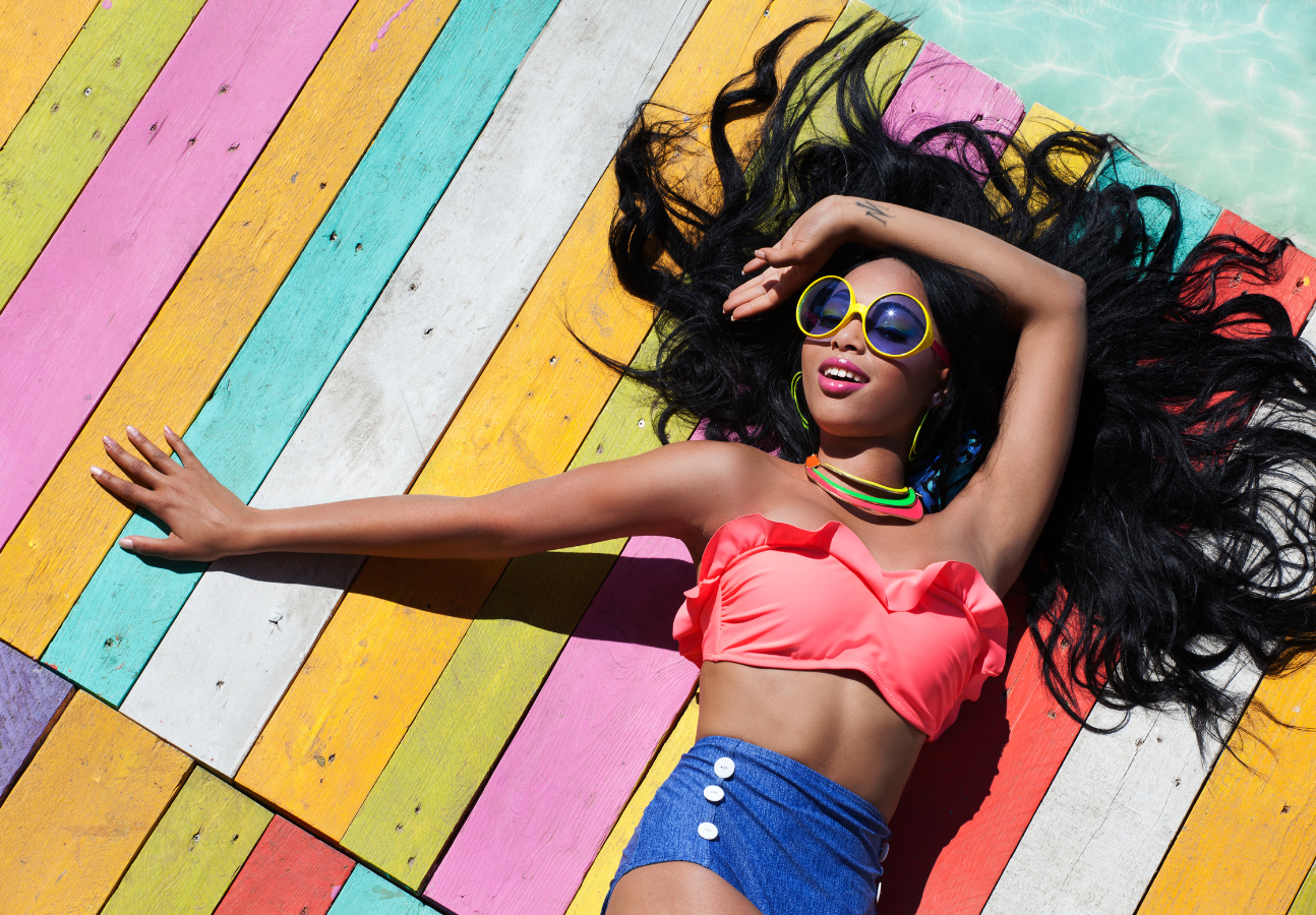 A black woman with dark hair and sunglasses in a two piece swimsuit lying down on a colorful background by the sea