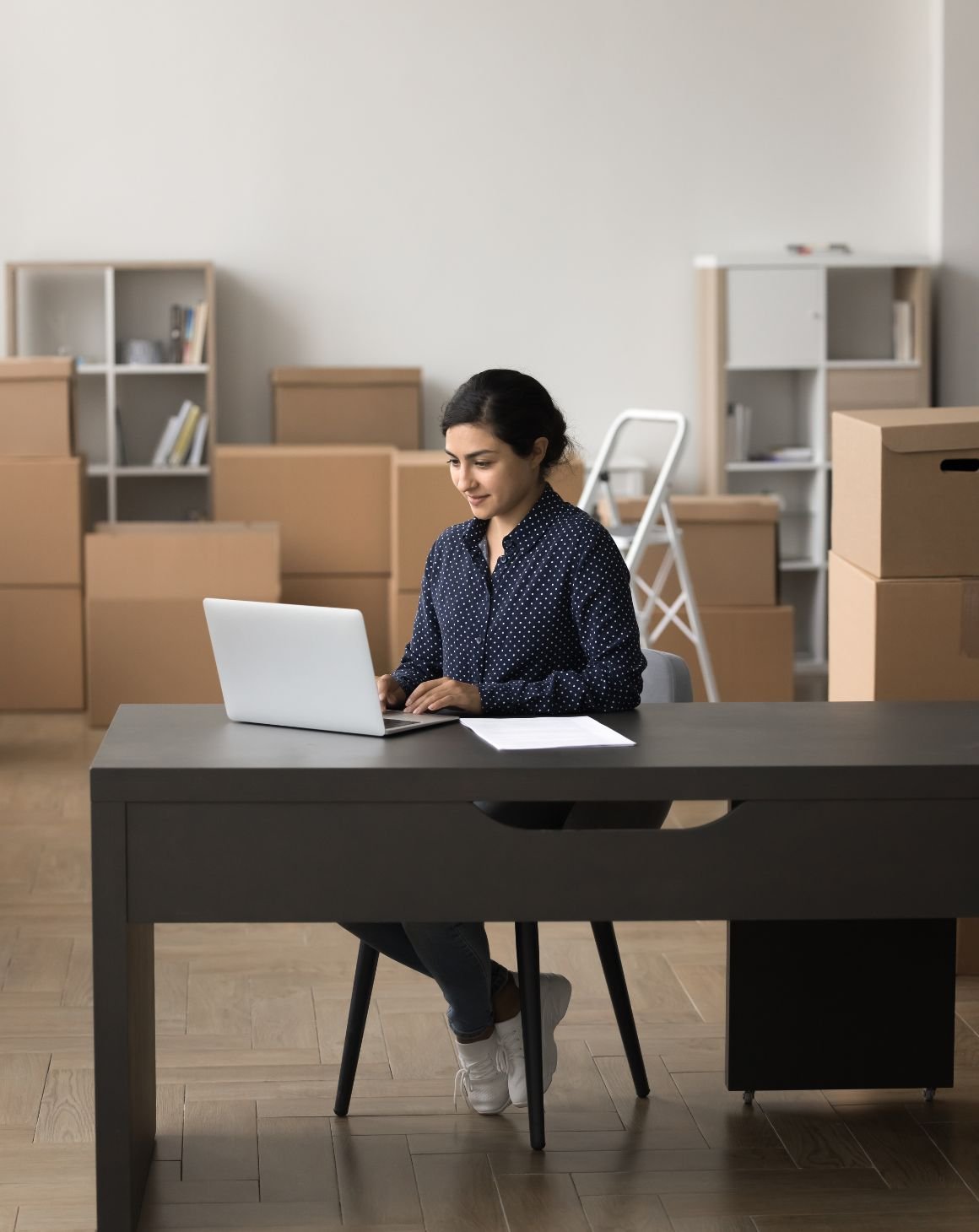 woman sitting on a desk on a laptop and boxes in the background