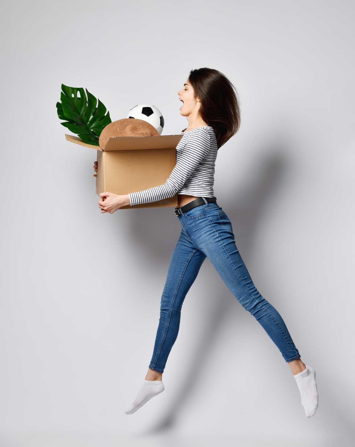 woman jumping and holding a cardboard box containing various items including a plant and a soccer ball