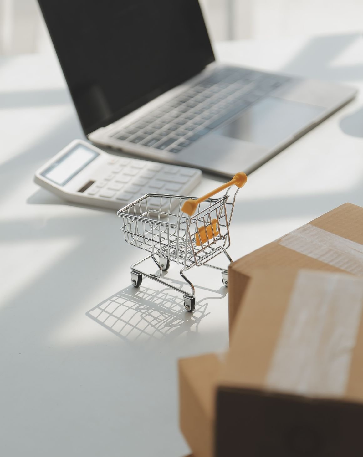 miniature shopping cart on a desk with a laptop, calculator, and cardboard boxes